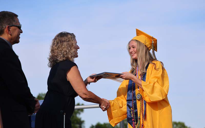 A man looks on as a woman hands a diploma to a young woman in a gold cap and gown. All are smiling.