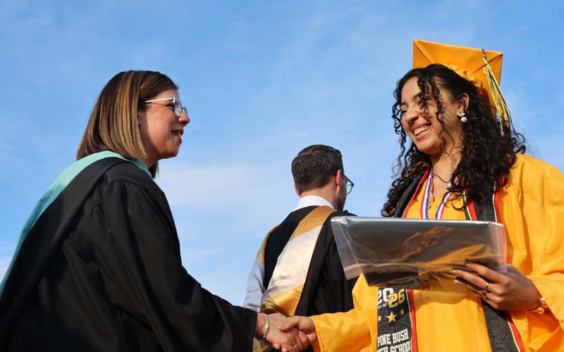 A woman shakes hands with a young woman in a gold cap and gown.