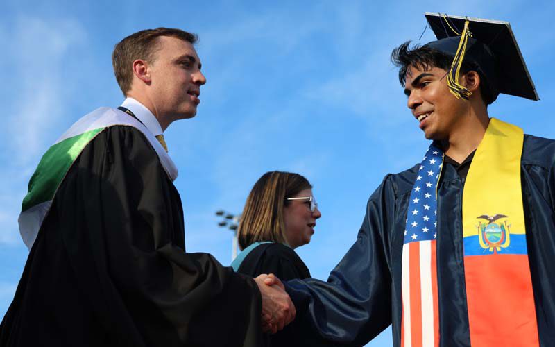 A man in a black graduation gown shakes the hand of a young man in a blue cap and gown.