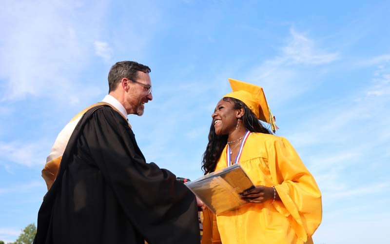 A girl in a gold cap and gown smiles and shakes the hand of a man in a black graduation gown.
