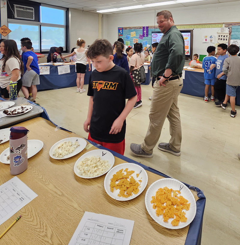 An elementary age boy stands at a table ready to taste from two different plates.