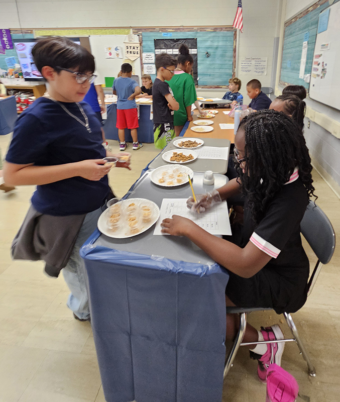 A fourth-grade girl sits at a table writing while a boy stands tasting from two plates of food.
