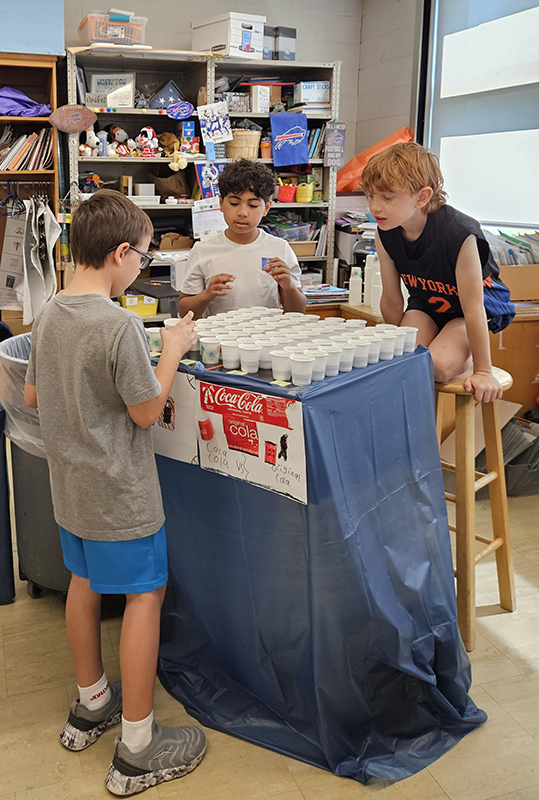 Two elementary age boys stand behind a table while another boy tastes soda.