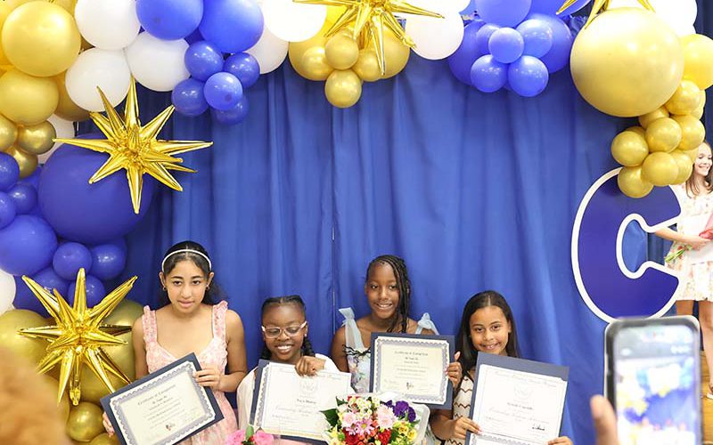 A blue background with blue gold and white balloons arched around it. There are four fifth-grade girls standing there holding certificates.