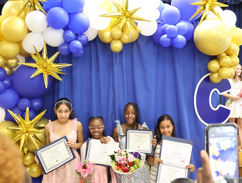 A blue background with blue gold and white balloons arched around it. There are four fifth-grade girls standing there holding certificates.