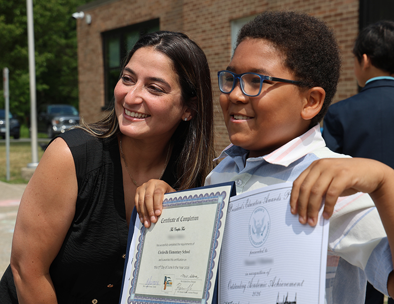 A woman leans in and smiles with a young man who is holding certificates.