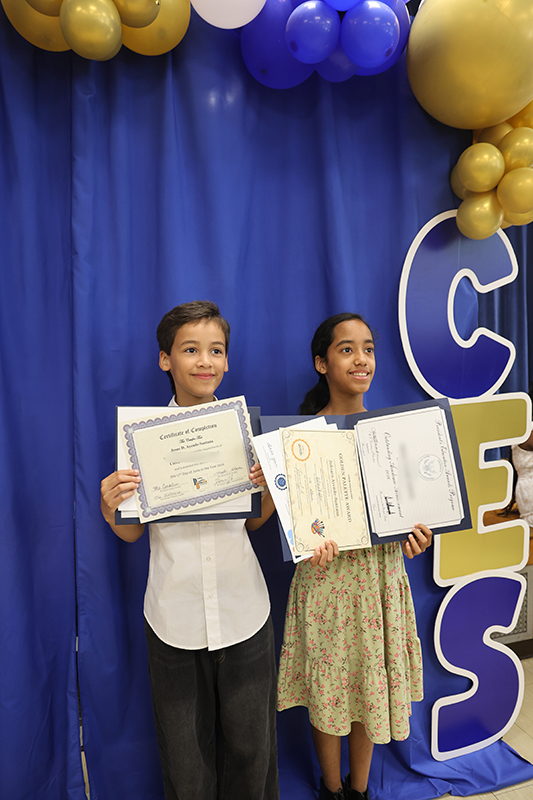 A fifth-grade boy and girl stand together smiling. They are holding certificates they earned. Behind them is a blue backdrop with CES on it and balloons over them.