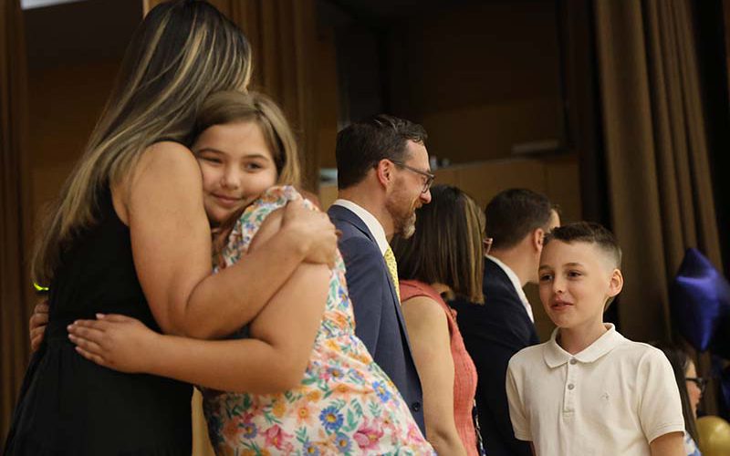 A fifth-grade girl hugs a woman on a stage. There are other adults behind them shaking hands with other kids.