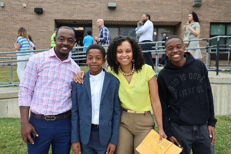 A family stands together outside a building. They are smiling. There is a man, a woman and two young men.