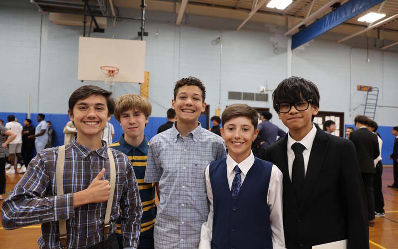 A group of five eighth-grade boys stand together inside a gymnasium smiling. They are all dressed nicely.