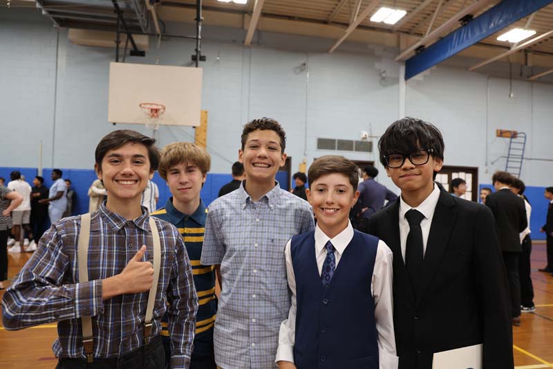 A group of five eighth-grade boys stand together inside a gymnasium smiling. They are all dressed nicely.