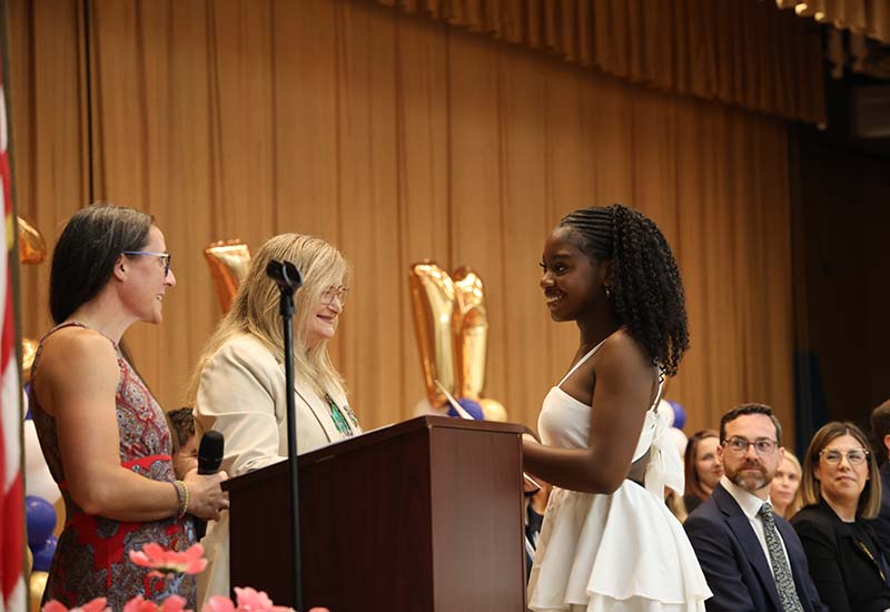 A woman with long blonde hair gives an award to an eighth-grade girl  as others look on.