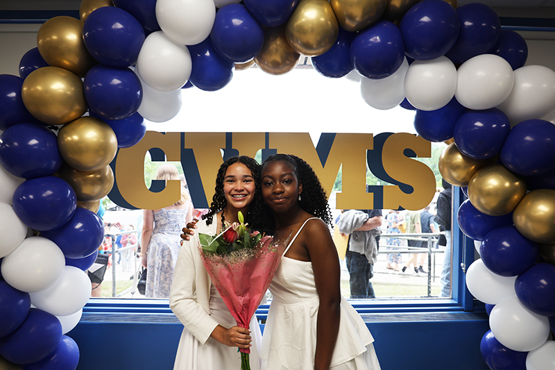 Blue, white and gold balloons in an arch. Two eighth-grade girls stand underneath it holding flowers and smiling.
