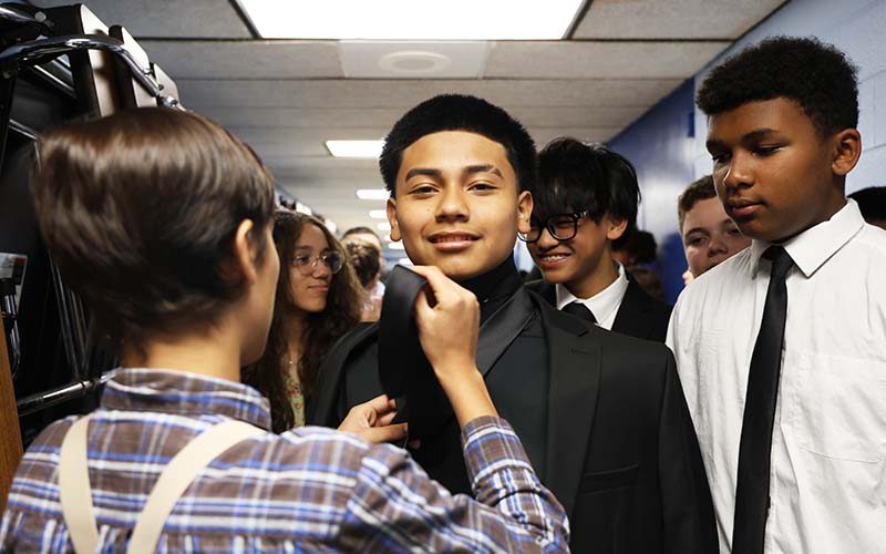 In a hallway of a school, a group of kids wait to begin their procession into their moving-up ceremony. One kid is tying the tie of another kid.