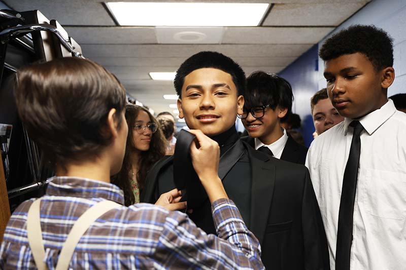 In a hallway of a school, a group of kids wait to begin their procession into their moving-up ceremony. One kid is tying the tie of another kid.