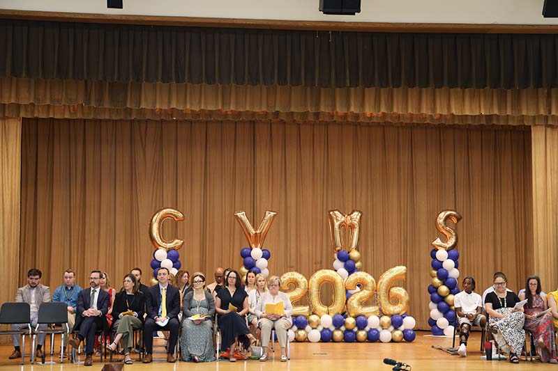 A photo of a stage with several adults sitting in chairs. There are gold balloons that say CVMS 2026.