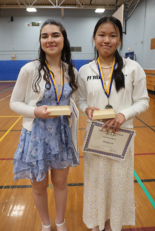 Two eighth-grade girls stand  together smiling. They both are wearing medals and holding certificates.
