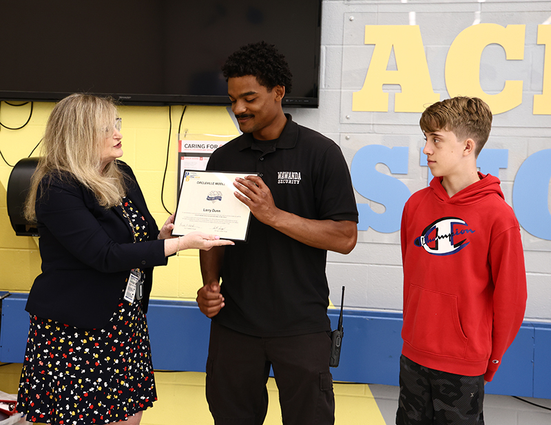 A woman presents a certificate to a young adult man who is smiling. A middle school age boy stands next to them.