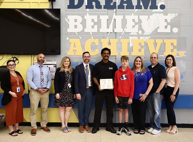 Eight adults and one middle school kid stand together. Behind them are large letters that say Dream Believe Achieve. The guy in the center holds a certificate.