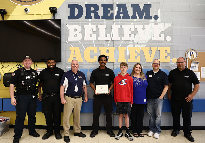 Seven adults and one middle school student stand together smiling. The man in the center holds a certificate. Behind them are letters that say Dream Believe Achieve.
