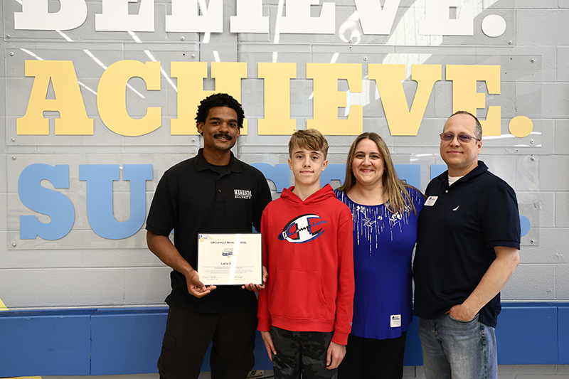 A young man holds a certificate and stands with a middle school kid and his mom and dad.