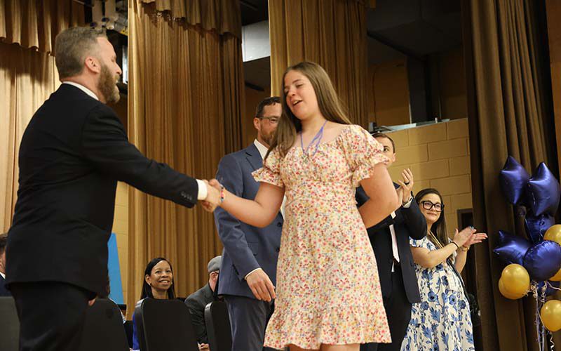 A fifth-grade girl shakes hands with a man on a stage.