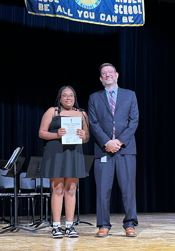 A man in a suit smiles and stands next to a middle school girl who is holding a certificate.
