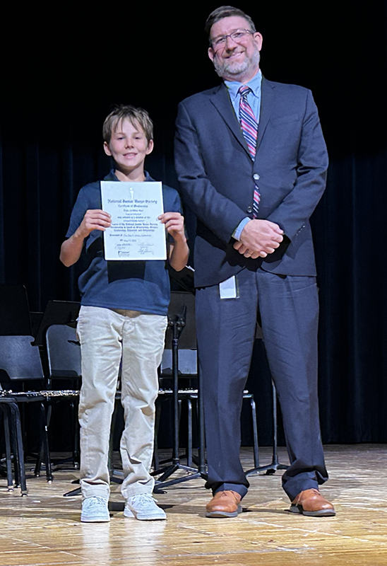 A man in a suit smiles as he stands next to a middle school kid who is holding a certificate.