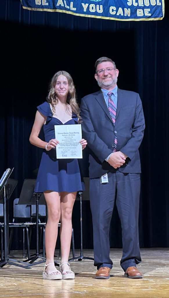 A man in a suit smiles as he stands next to a middle school kid who is holding a certificate.