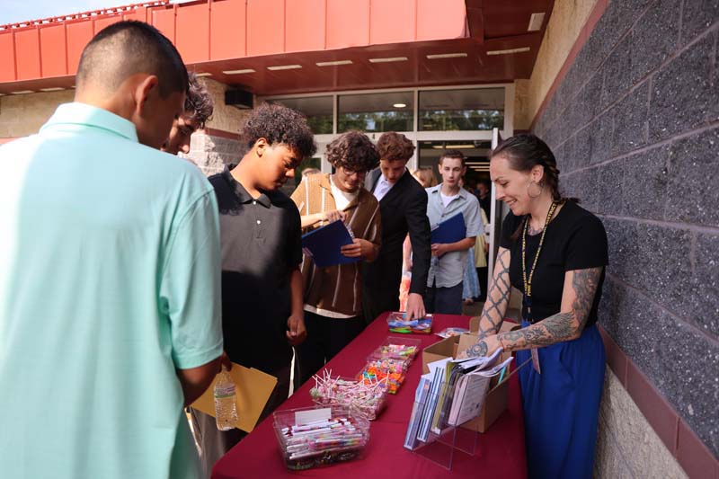 A woman stands at a table filled with little goodies for kids to take, like pencils and erasers. There are several middle school kids around the table.