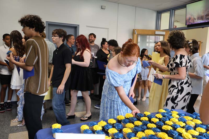 A hallway filled with middle school kids. There is a table with blue and gold cupcakes. kids are taking cupcakes from the table.