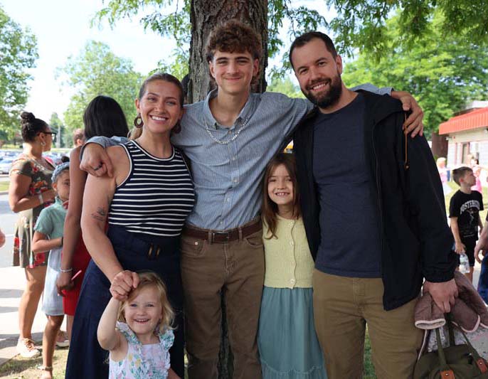 A young man has his arms around a man and a woman. There are two little girls there too. All are smiling.