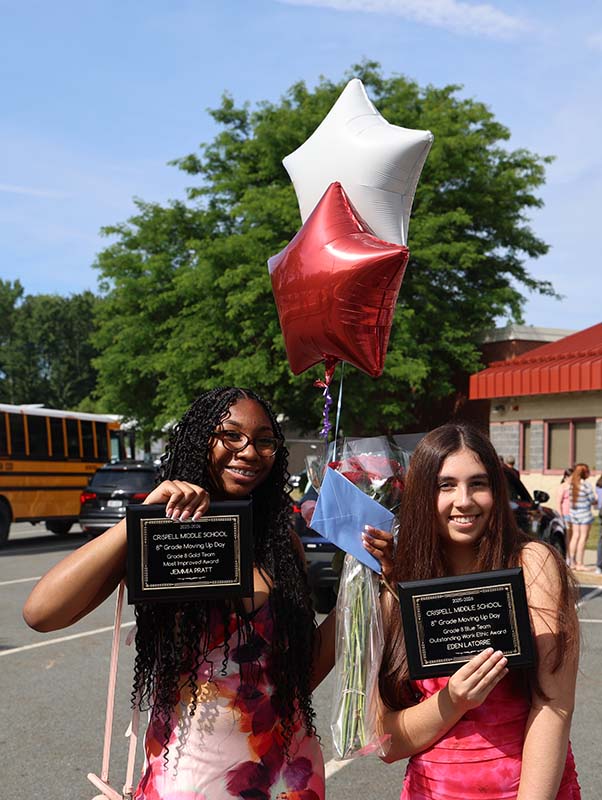 Two eighth-grade girls stand outside of a school building on a bright sunny day. They are smiling and holding balloons and awards.