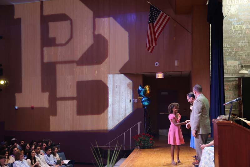 An eighth-grade girl in a pink dress shakes hands with adult as she walks across a stage. There is a large PB reflected on the wall behind her.
