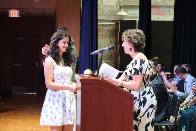 A young woman in a printed dress smiles as she accepts an award from a woman behind a podium.