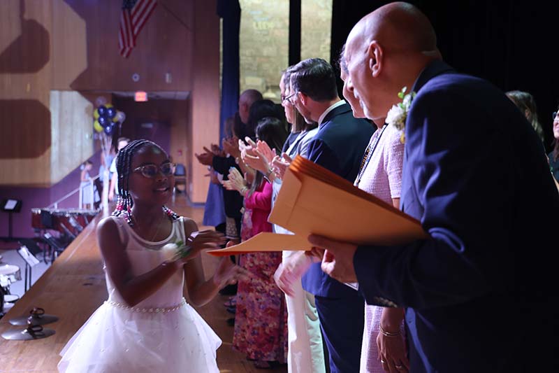 A fifth-grade girl dressed in a white dress shakes hands with adults.