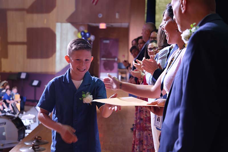 A fifth-grade boy shakes hands with adults on a stage.