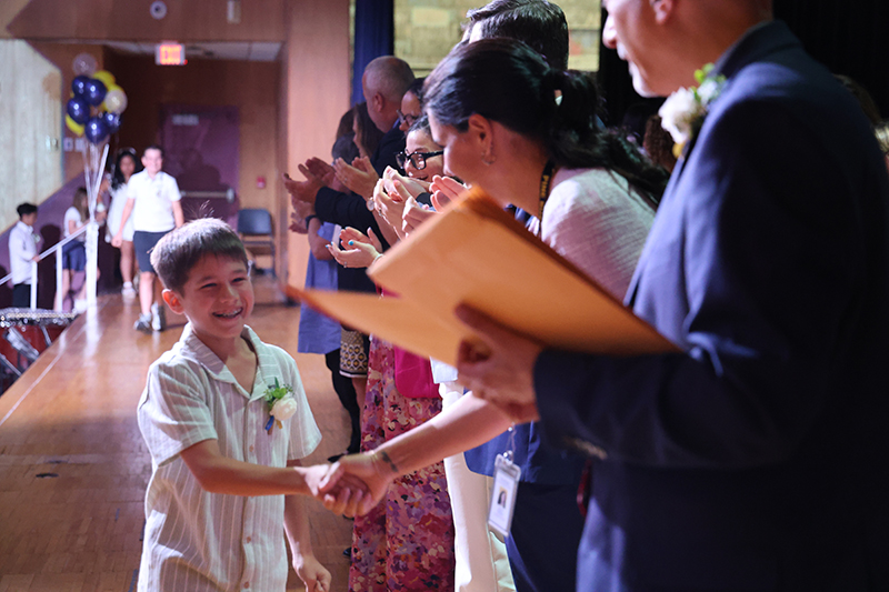 A fifth-grade boy smiles as he shakes hands with a woman. A man stands next to them with envelopes to hand out. Others are clapping.