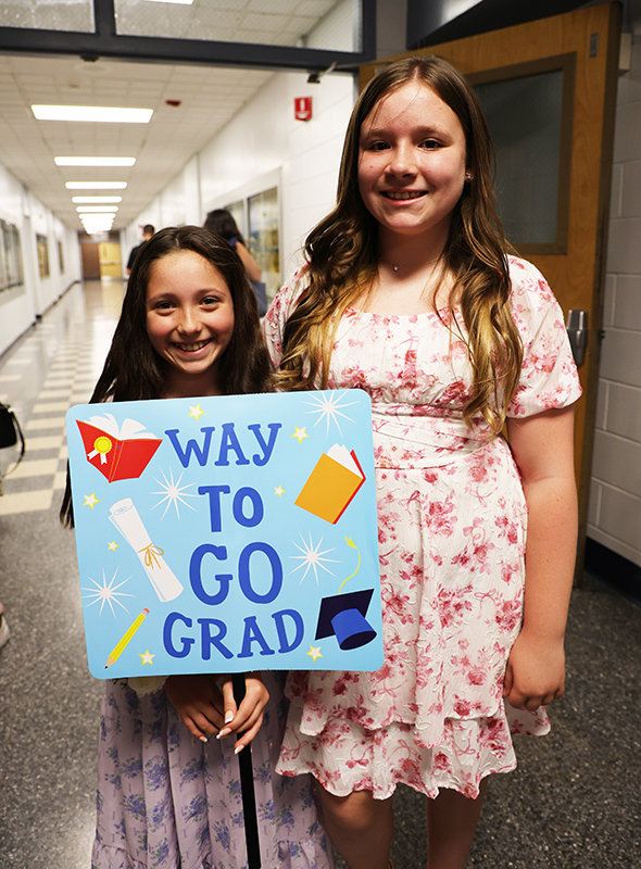 Two fifth-grade girls stand together smiling. One girl has a sign that says Way to go grad!