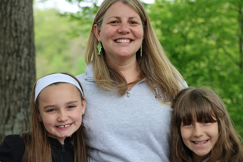 A woman with long blonde hair smiles wearing trout earrings. On either side is a little girl also smiling.