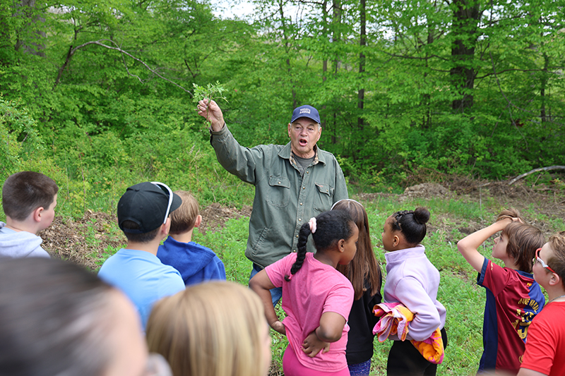 A man holds up a leaf while talking to a group of third-grade students.