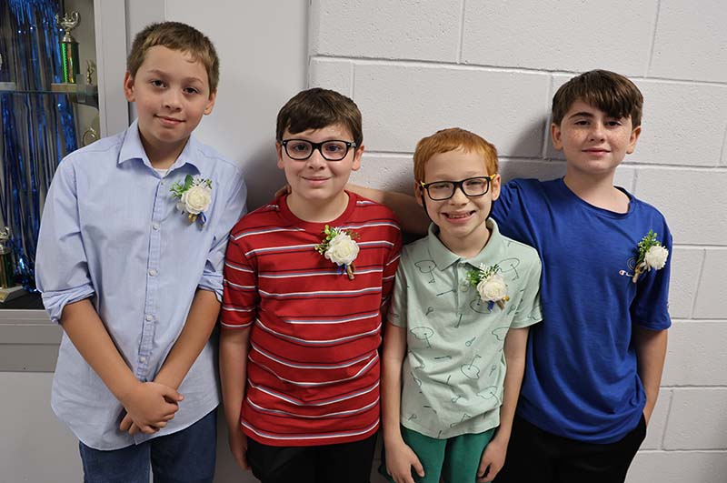 Four boys stand together. They each have a flower pinned to their shirts. and are smiling.