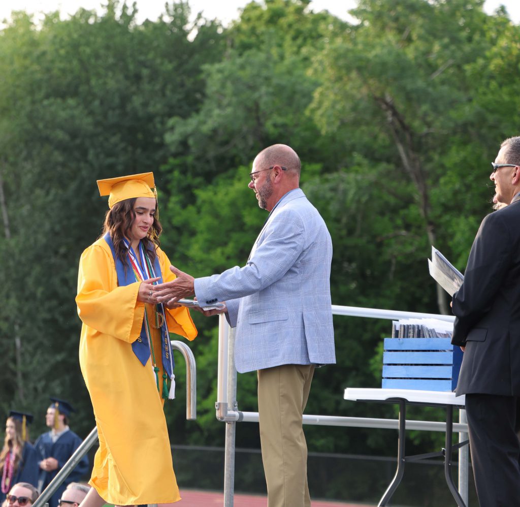 A man in a light blue jacket hands a diploma to a young woman in a gold cap and gown.