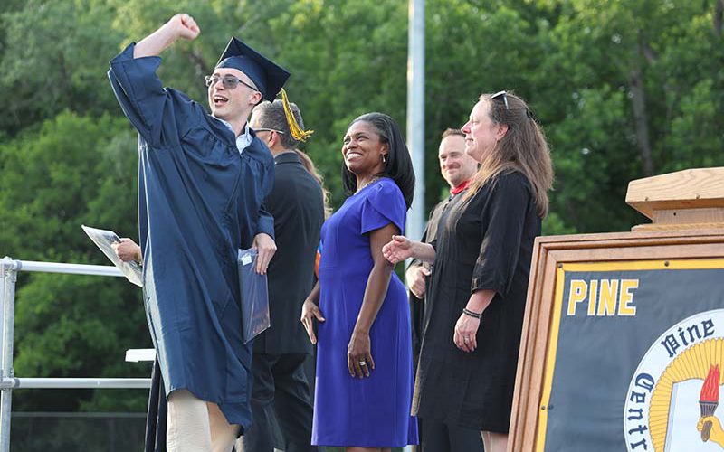 A young man in a navy blue cap and gown pumps his fist into the air as he walks across a stage to get his diploma. Adults are looking on smiling.