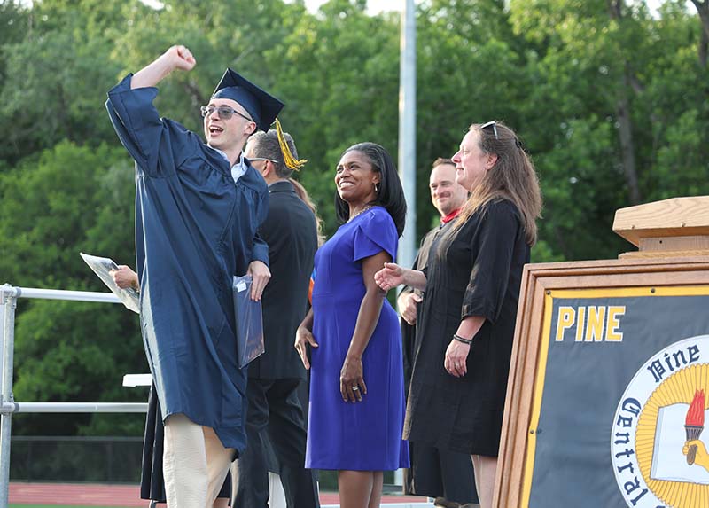 A young man in a navy blue cap and gown pumps his fist into the air as he walks across a stage to get his diploma. Adults are looking on smiling.