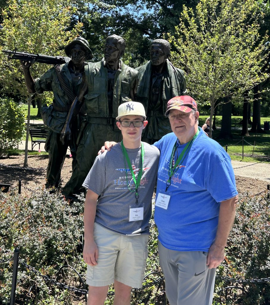 A man in a blue shirt and baseball cap stands with a young man with glasses and a baseball cap in front of a statue.
