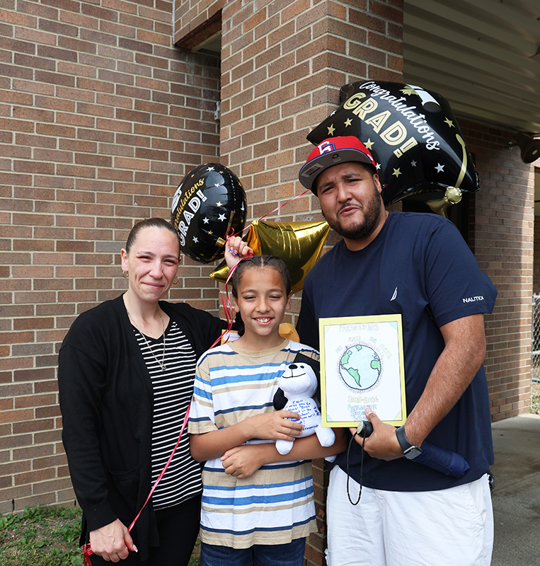 A man on the right and a woman on the left hold balloons and a yearbook. There is a fifth-grade kid in the center.