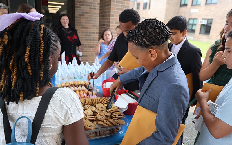 A group of fifth-grade kids gather around a table and tke cookies.