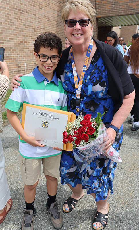 A woman wearing a blue dress and sunglasses holds flowers and has her arm around a fifth-grade boy who is also smiling.