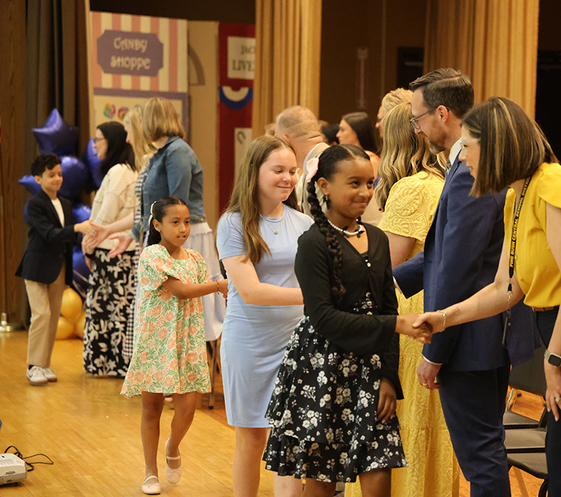 A line of fifth-grade girls shake hands with adults on a stage.